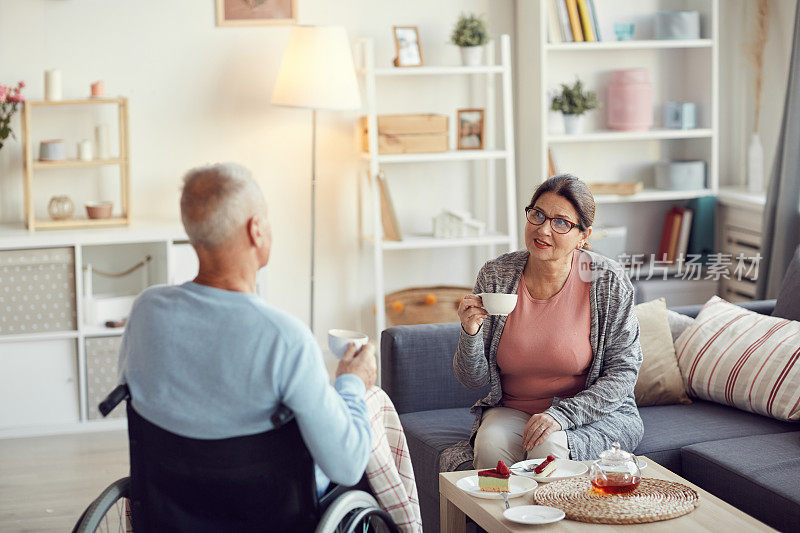 Content senior woman in家装坐在沙发上，一边喝茶一边微笑着与残疾的丈夫交流
